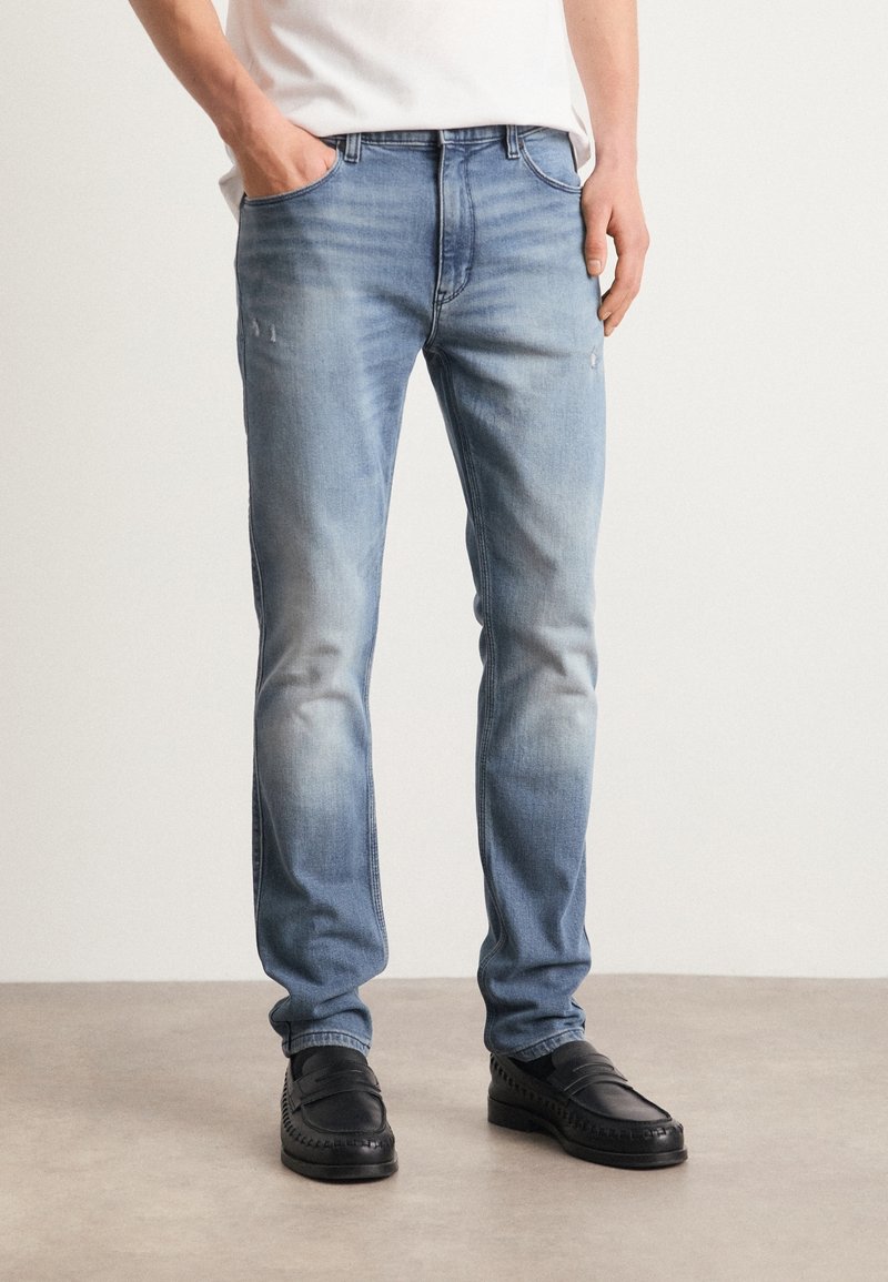 Homme portant un jean slim bleu clair avec délavé et petits détails usés, mocassins noirs et chemise blanche rentrée.