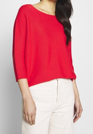 Red knitted sweater with a wide neckline and three-quarter sleeves, paired with light-colored trousers. Smooth texture, casual fit.