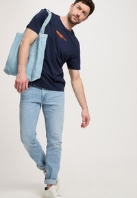 Granatowa bawełniana koszulka z pomarańczowym logo, jasnoniebieskie dżinsy i białe sneakersy. Jasnoniebieska torba na ramię zawieszona na ramieniu.