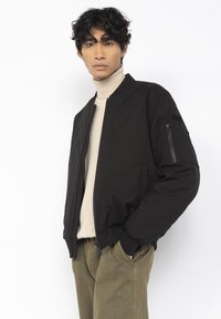 Schwarze Bomberjacke mit einer mit Reißverschluss versehenen Tasche am linken Ärmel, leichtem Stoff, gerippten Bündchen und kragenlosem Design.