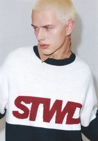 Młoda osoba z krótkimi, rozjaśnionymi włosami, ubrana w biały sweter z czerwonym napisem "SNW" oraz brązową koszulę w kratę z kołnierzykiem pod spodem.