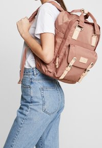 Vaaleanpunainen nylon-reppu, jossa on kermanväriset hihnat, yläkahvat ja etutasku. Ominaisuuksina suorakulmainen muoto ja ompeluakkentit. Kaksi sivutaskua.