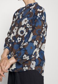 Blusa floreale con colori blu, marrone e bianco. Realizzata in tessuto leggero, con scollatura rotonda e maniche con bottoni.