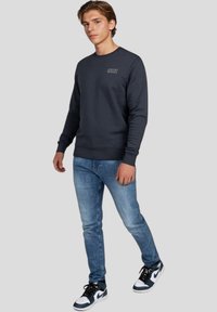 Tmavě šedá mikina s kulatým výstřihem a logem, spárovaná se světle modrými džíny a černobílými teniskami. Neformální, pohodlný outfit.