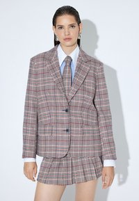 Geruite pak met een op maat gemaakte blazer en een geplooide mini-rok in tinten van rood, groen en beige; bevat een blauwe das en een wit overhemd eronder.