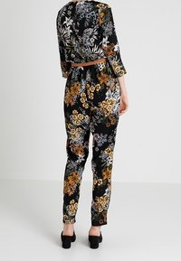 Salopette floreale in tessuto nero con motivi gialli, arancioni e bianchi. Presenta maniche a tre quarti e una vita accentuata da una cintura.