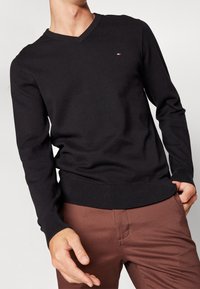 Uomo che indossa un maglione nero a V con un piccolo logo Tommy Hilfiger e pantaloni marroni, una mano in tasca, l'altra rilassata di fianco.