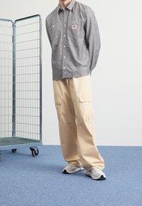 Man draagt een grijs overhemd met knopen, beige cargobroek en witte sneakers, staat op een blauw tapijt naast een metalen verrijdbaar rek.