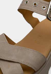 Κοντινό πλάνο μπεζ σουέντ σανδάλι με σταυρωτούς ιμάντες, μεταλλική αγκράφα και μαλακό ταμπά πέλμα.