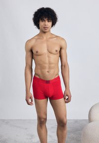 Boxer briefs vermelhos de algodão com um ajuste justo, apresentando uma cintura elástica preta com marca. O modelo tem cabelo encaracolado e um físico tonificado, em pé sobre uma superfície cinza claro.