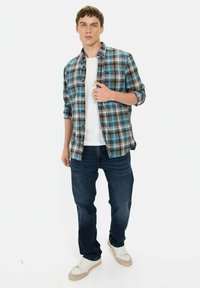 Blauw en zwart geruit overhemd over een wit T-shirt, gecombineerd met donkere jeans en witte sneakers, staand voor een effen achtergrond.