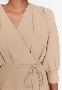 Beige zabalovací šaty s véčkovým výstřihem, skladovanými rukávy a opaskem na zavázání. Tkanina má hladkou texturu a mírný lesk.