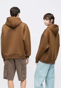 Rudi gobtuvių džemperiai su ribotais apatiniais kraštais, dėvimi su pilkomis kargo šortais ir šviesiai mėlynais plačiomis kojų kelnėmis, rodomi iš nugaros ir šono.