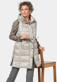 Helles graues gepolstertes Weste mit Kapuze, mit Druckknöpfen und einem gesteppten Design. Getragen über einem gemusterten Pullover und karierter Hose.