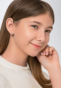 Pendientes de botón de unicornio plateados con detalles de colores. El modelo tiene el cabello castaño liso y lleva una camisa blanca de cuello alto.