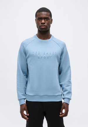 Άνδρας που φοράει ένα ανοιχτό μπλε φούτερ με την ανάγλυφη επιγραφή «TED BAKER SPORT» μπροστά, στέκεται μπροστά σε ένα απλό λευκό φόντο.