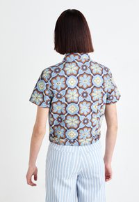 Kortermet skjorte i lyseblått stoff med et blomsterdesign i gult, brunt og blått; cropped design med knapppdetaljer bak.
