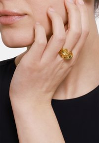 Goldene, gedrehte Ring mit polierter Oberfläche, zwischen den Fingern gehalten. Glatte Textur und skulpturaler Entwurf, vor einem dunklen Hintergrund.