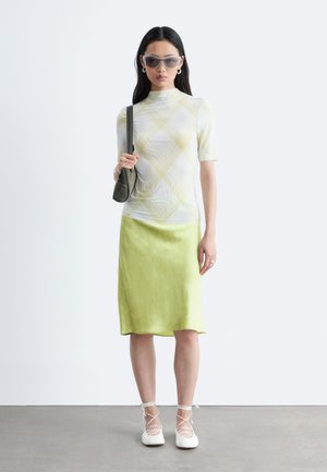 Vrouw met grijze zonnebril, wit-geel geruit topje met korte mouwen, lichtgroene satijnen rok, witte veterschoenen en een grijze schoudertas.