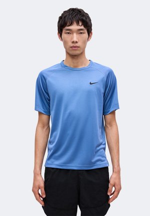 Jeune homme aux cheveux courts et foncés portant un t-shirt de sport bleu Nike à manches courtes et un short noir, debout devant un fond uni.