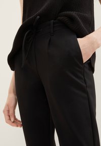 Černé elastické kalhoty se šňůrkou v pase, bočními kapsami a hladkým povrchem, částečně pokryté pleteným tmavým topem.