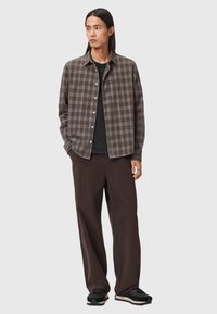 Karierter Shirt in Braun, Grau und Creme, geknöpft, mit einem klassischen Kragen über einem schwarzen Shirt. Kombiniert mit dunklen braunen weiten Hosen und schwarzen Turnschuhen.