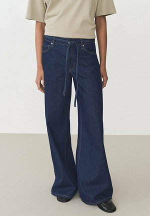 Wijde denimjeans in een donkerblauwe kleur, hoge taille en verstelbare trekkoord. Gecombineerd met een beige oversized t-shirt en zwarte schoenen.