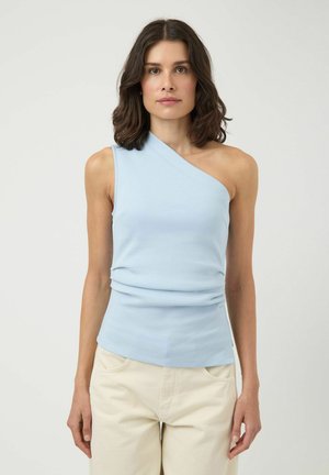 Femme aux cheveux châtain foncé mi-longs portant un haut bleu clair à une épaule et un pantalon beige, debout devant un fond uni.