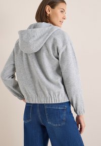 Sudadera gris con capucha y cordones, puños acanalados y dobladillo. Combinada con jeans de mezclilla azul. Textura suave, corte casual y diseño relajado.