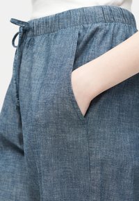 Main dans la poche d'un pantalon bleu chambray à cordon de serrage avec taille élastique, porté avec un haut blanc.