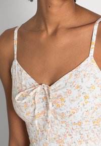 Top de estampado floral con tejido ligero, tirantes finos ajustables y un detalle de lazo en el escote. Los colores incluyen blanco, amarillo y naranja.