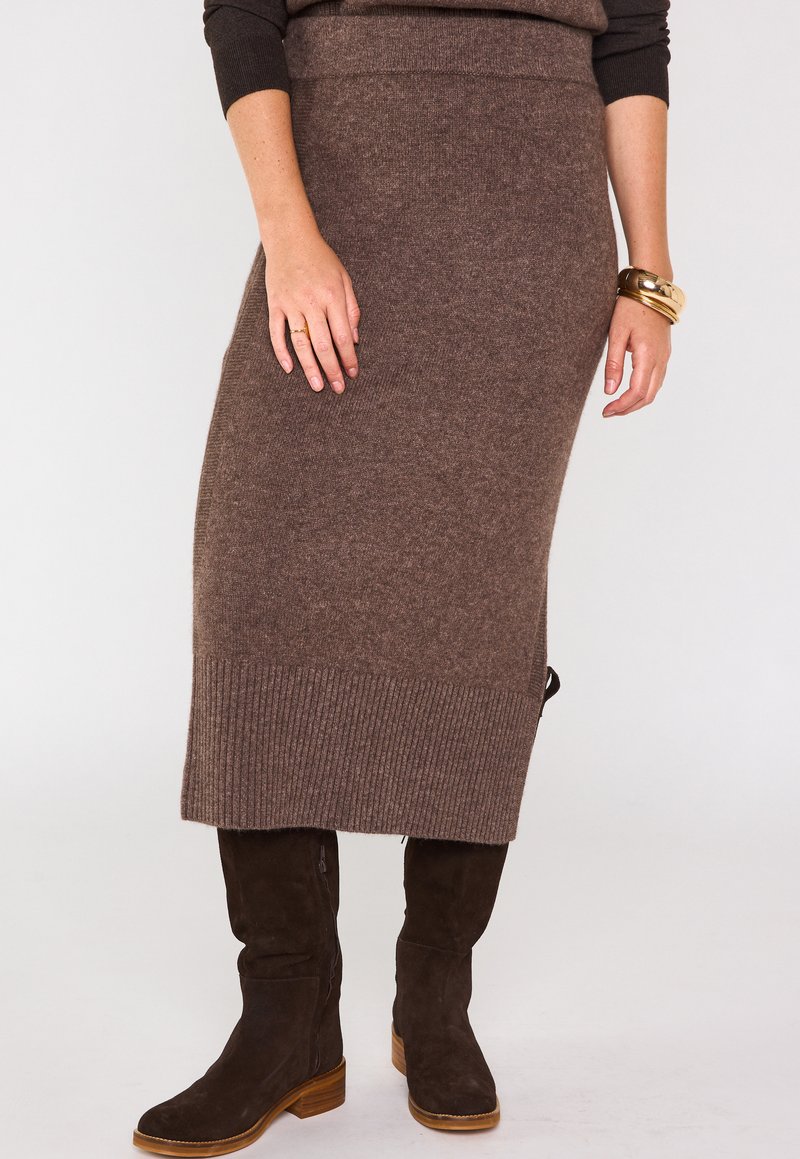 Jupe midi en tricot marron avec un ourlet côtelé, taille ajustée et détail sur le côté. Associée à des bottes en suède marron jusqu'aux genoux et un bracelet en or.