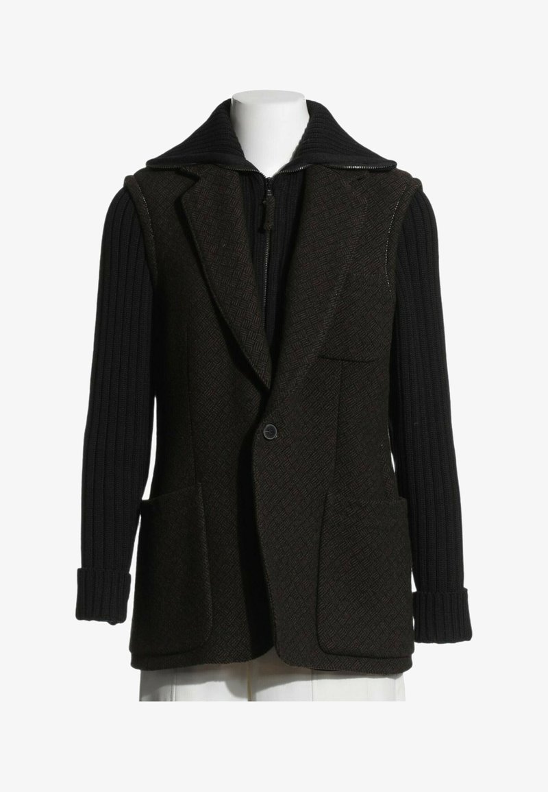 Gilet brun foncé texturé avec un bouton, grandes poches avant, porté sur des manches longues noires côtelées avec un col zippé.