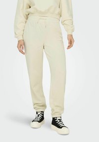 Beige Jogginghosen mit elastischem Bund und Bündchen, aus weichem Stoff. Kombiniert mit schwarzen High-Top-Sneakern mit weißen Schnürsenkeln und Sohle.