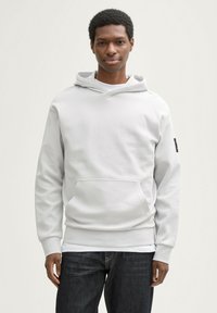 Felpa grigia chiara con tasca a marsupio, cappuccio con cordino e polsini a coste. Indossata sopra jeans scuri, con un design di tessuto testurizzato.
