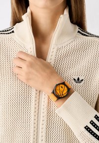 Reloj de pulsera negro y naranja con un diseño de baloncesto, que cuenta con una correa texturizada naranja y una caja redonda, usado con un suéter de punto crema con cremallera.