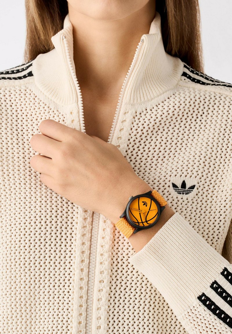 Reloj de pulsera negro y naranja con un diseño de baloncesto, que cuenta con una correa texturizada naranja y una caja redonda, usado con un suéter de punto crema con cremallera.