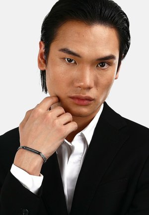 Svart jacka och vit skjorta; manlig modell med slickat hår. Tunn silverarmband på handleden; allvarligt uttryck, ljus hudton.