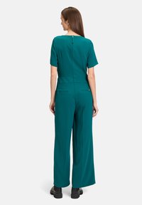 Teal jumpsuit met korte mouwen, wijde pijpen en een ritssluiting op de rug. Heeft twee achterzakken en een gladde, gestructureerde stof.