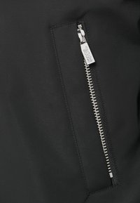 Μαύρο υφασμάτινο μπουφάν με τσέπη με φερμουάρ από μέταλλο. Υφή με κομψό, ματ φινίρισμα και διακριτικές λεπτομέρειες ραφής.