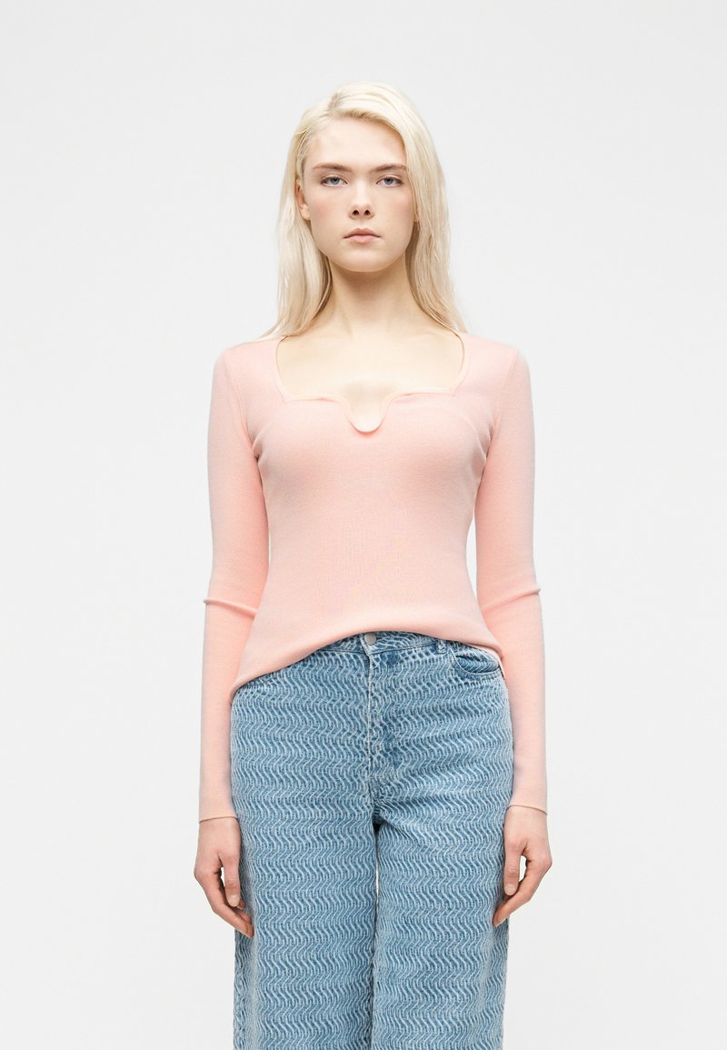 Ξανθιά γυναίκα που φοράει εφαρμοστή ανοιχτό ροζ μπλούζα με μακρύ μανίκι και ντεκολτέ με σχισμή, καθώς και ψηλόμεσα μπλε τζιν με σχέδιο, όρθια.