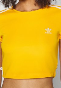 Žluté zkrácené tričko vyrobené z hladkého materiálu, s bílými pruhy na ramenech a vyšitým logem Adidas na hrudi.