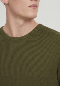 Olive roheline kudumitega sveter, millel on sooniline tekstuur, ümar kaelus ja lühikesed varrukad, rõhutades lõdva disaini.