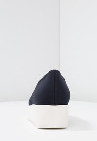 Marineblauwe zachte schoen van materiaal met een dikke witte zool, voorzien van een ronde achterkant en minimale stikseldetails. Glad oppervlak zonder versieringen.