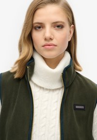 Ciemnozielona kamizelka polarowa z wysokim kołnierzem, złotym zamkiem i niebieskim akcentem. Noszona na białym golfie z dzianiny w warkocze. Logo na kamizelce.