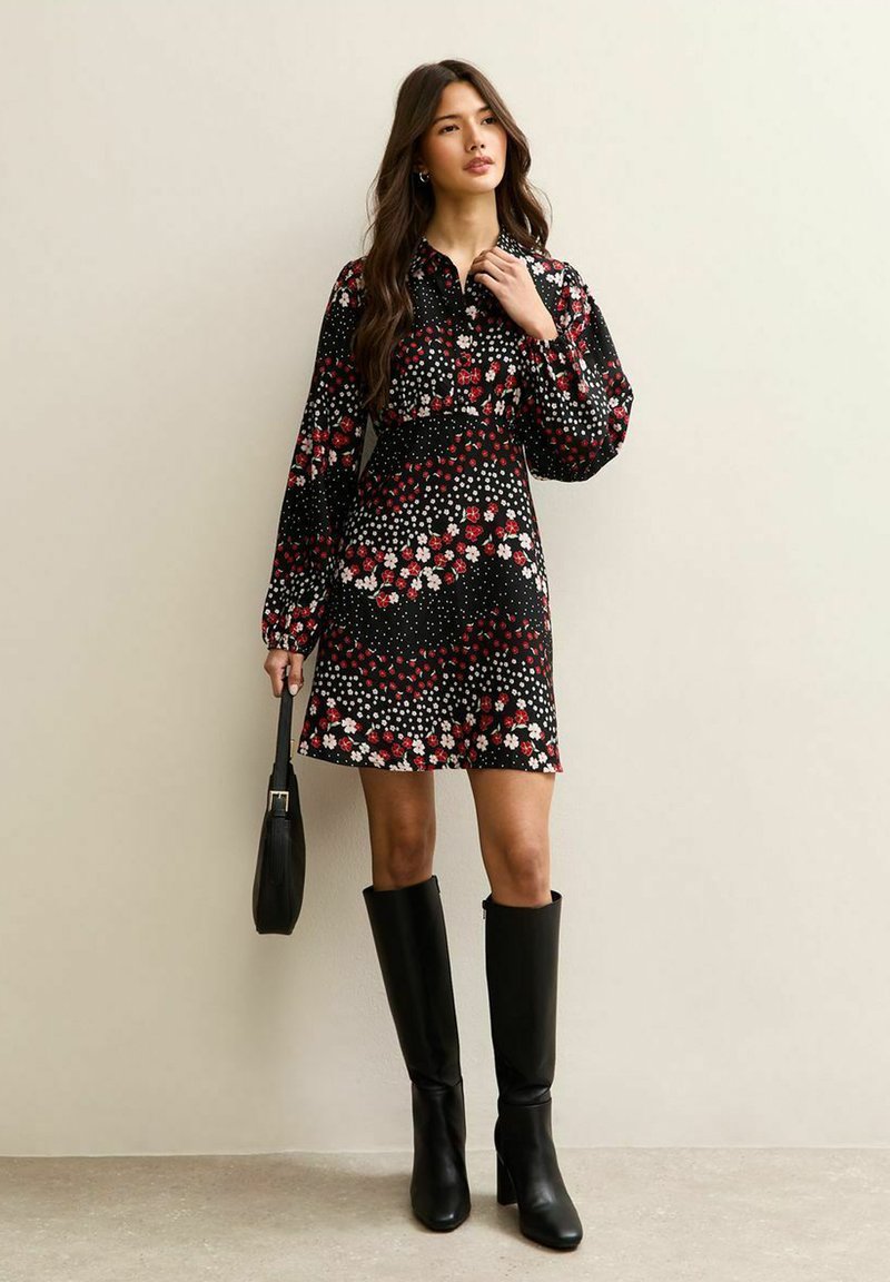 Vrouw in een zwarte bloemenjurk met lange mouwen, die een zwarte handtas vasthoudt en zwarte leren laarzen tot aan de knie draagt.