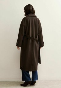 Bruine wollen trenchcoat met een ceintuur in de taille, een grote kraag en met knopen gesloten manchetten, gedragen over wijde jeans en hoge hakken.