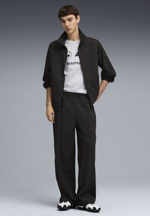 Giovane uomo con giacca e pantaloni da ginnastica neri, maglietta bianca BMW Motorsport e sneakers bianche e nere, in piedi davanti a uno sfondo grigio.