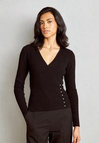 Haut noir à manches longues côtelé avec un col en V et des boutons décoratifs sur le côté gauche. Le tissu a une texture lisse.