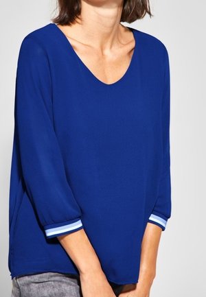 Femme portant un chemisier bleu roi à manches longues avec des poignets rayés blancs et bleu clair, associé à un pantalon gris.